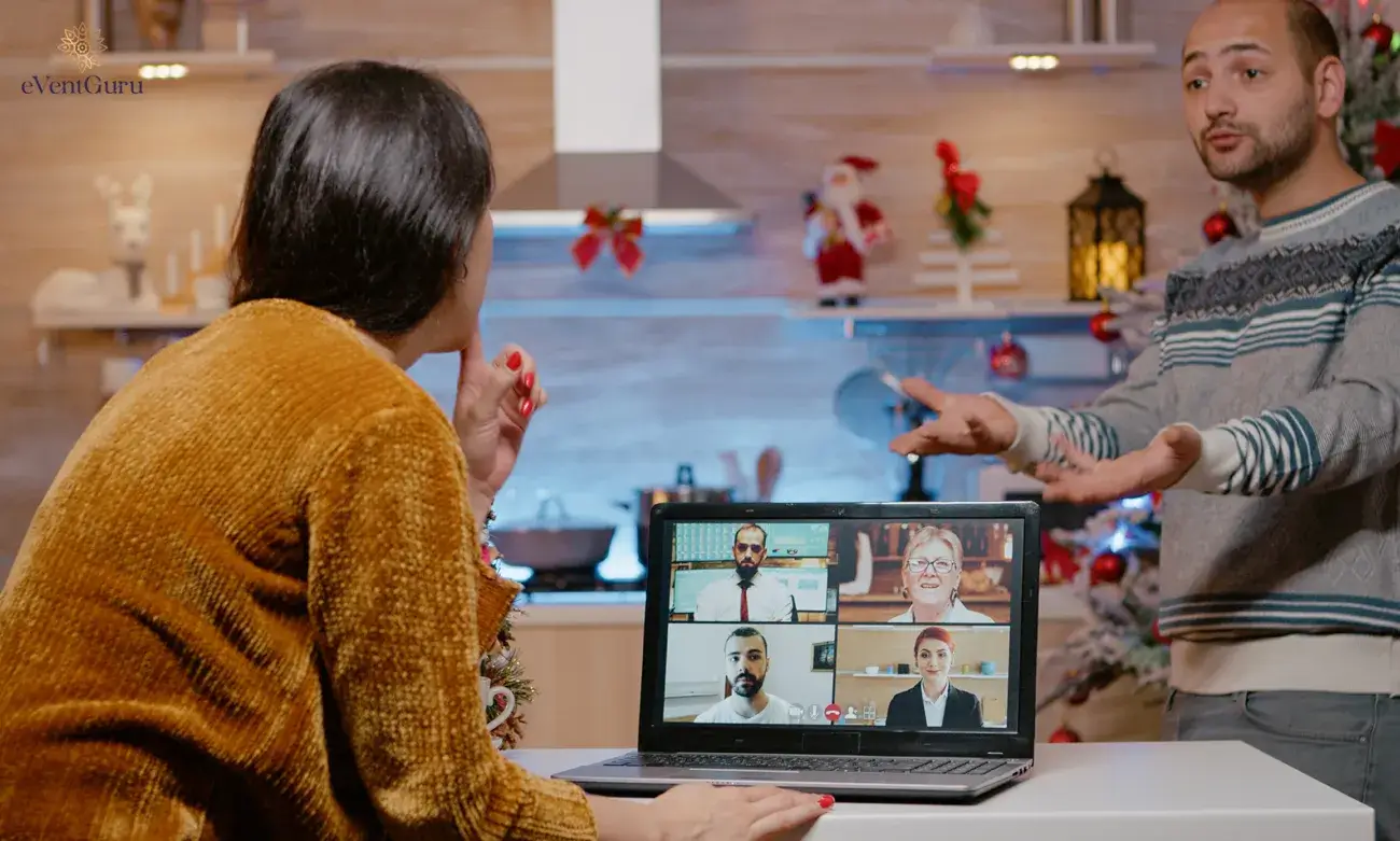 Two people engaged in a video call on a laptop with four colleagues, set in a festively decorated kitchen. Holiday decorations, including ornaments and small Santa figurines, are visible in the background.